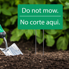 Load image into Gallery viewer, National Etching Do not mow bilingual green acrylic garden sign shown staked into earth. Green leaves are blurred in background and a hand trowel is shown to the left of sign